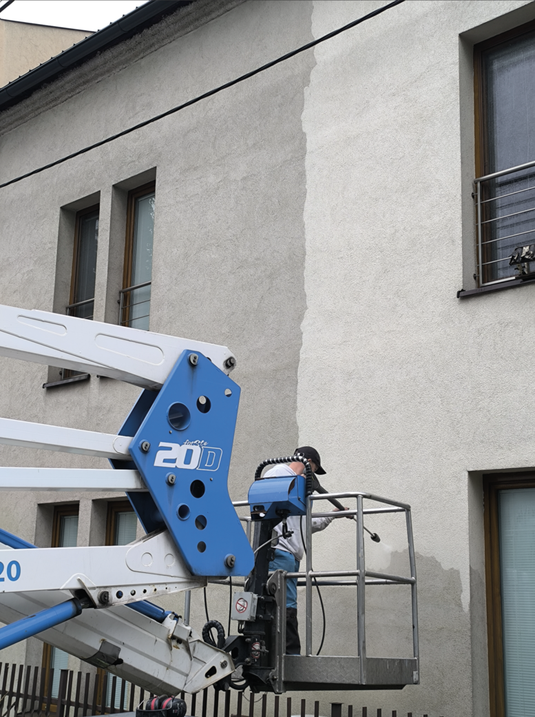 Worker on a blue and white lift cleaning the façade of a building with a pressure washer, showing a visible difference between cleaned and uncleaned wall sections.