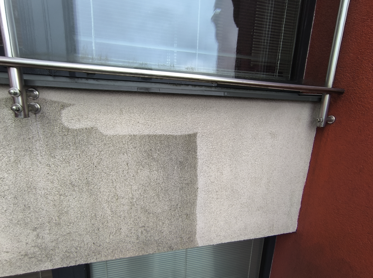 Section of an outer building wall showing a clear contrast between a cleaned light gray concrete area and a darker dirty section, with a red wall and metal railing above.
