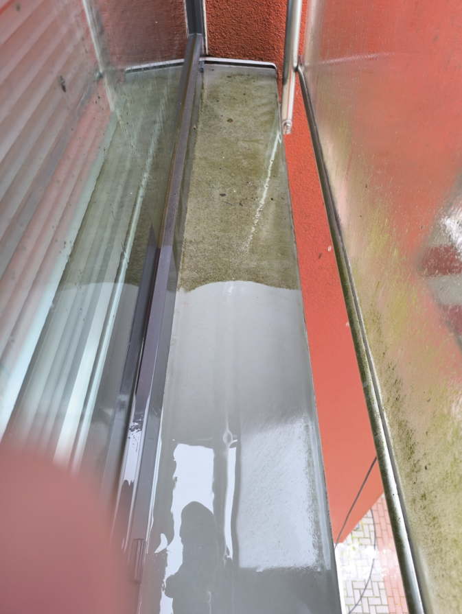 A narrow balcony with a wet, partially cleaned surface, reflecting the adjacent orange wall and glass window.