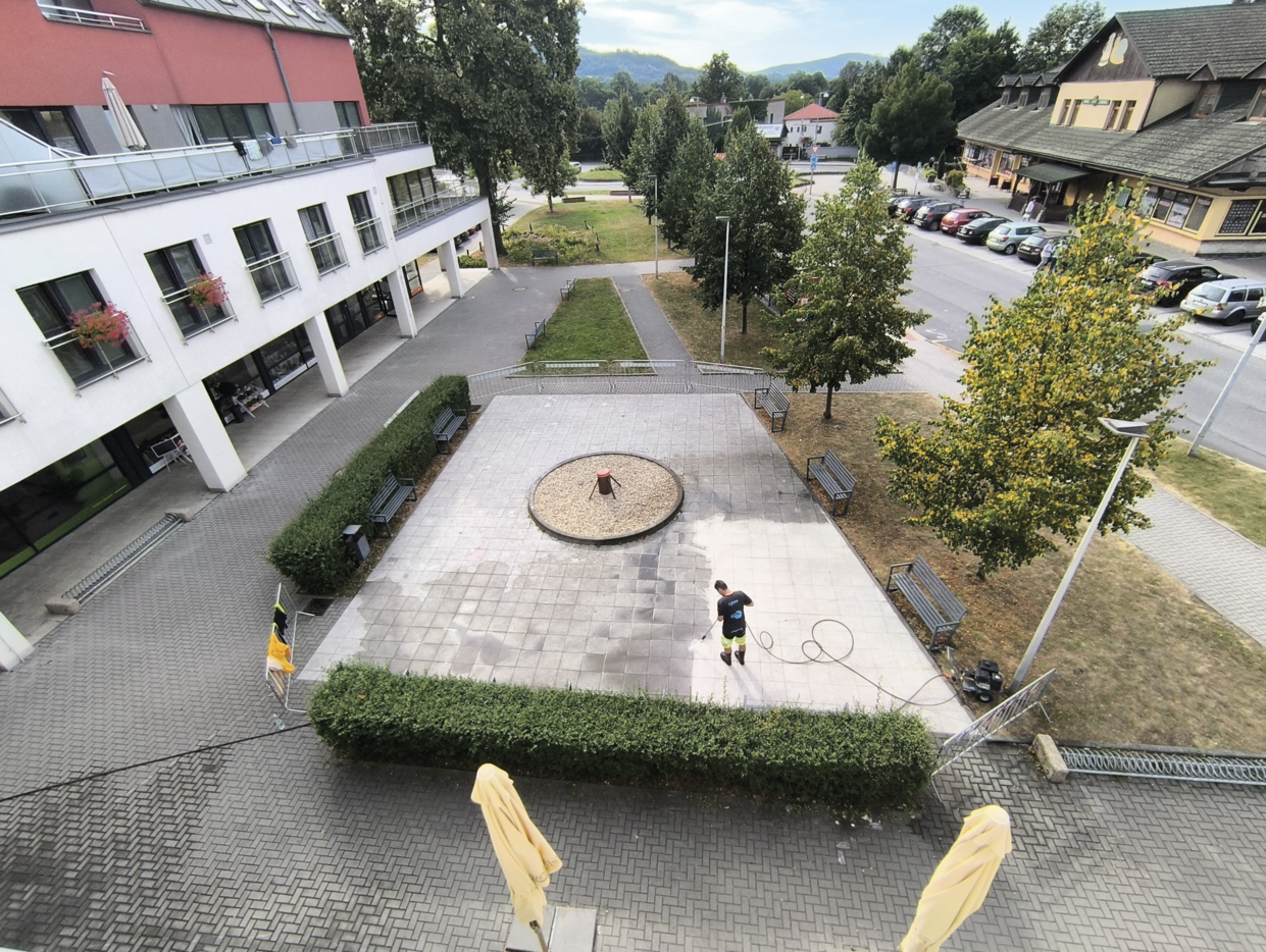 Person power washing a large tiled courtyard with benches and trees surrounding it.