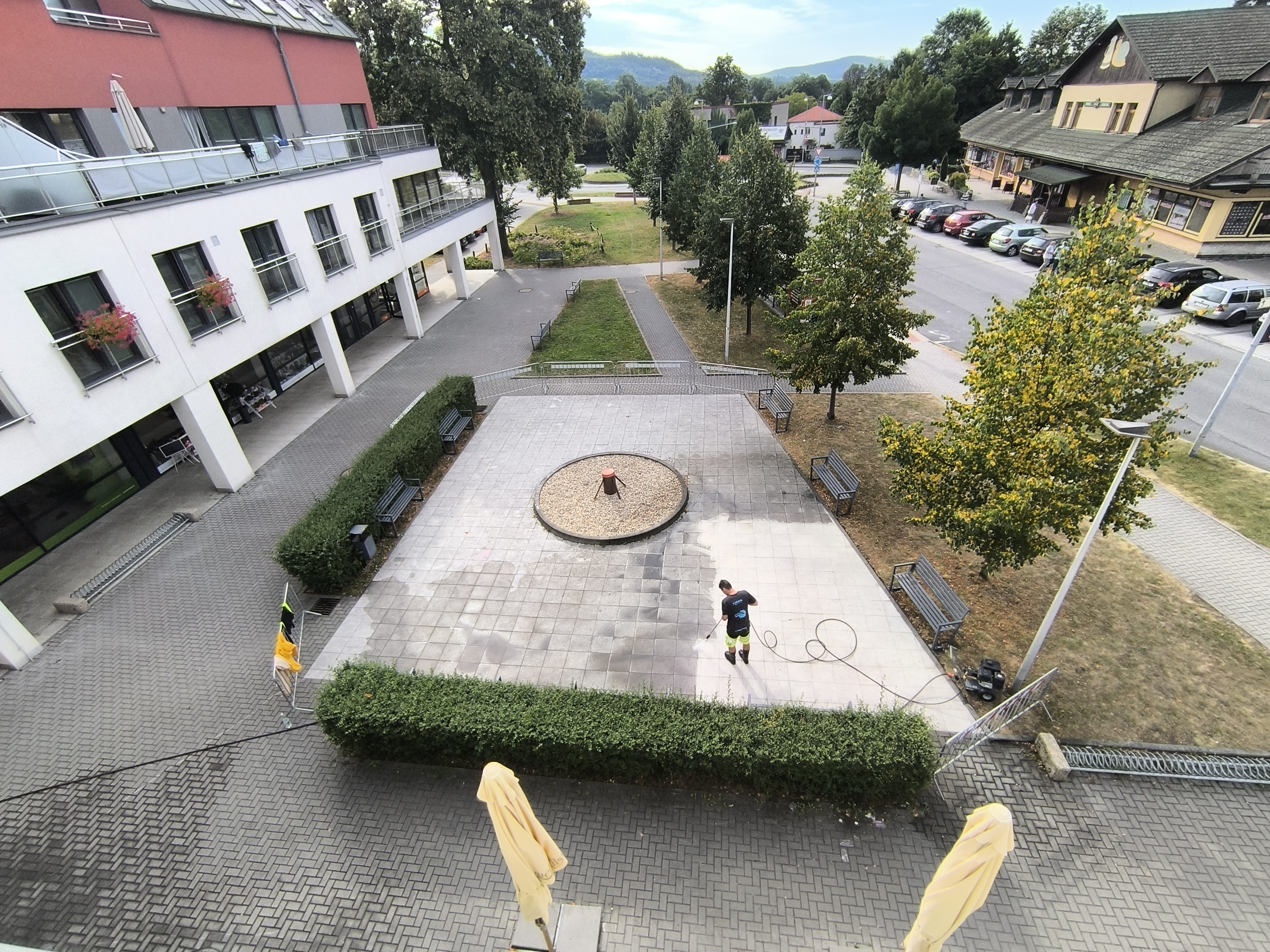 Person pressure washing a large paved patio area with benches and trees around, adjacent to a white building and a street with parked cars.