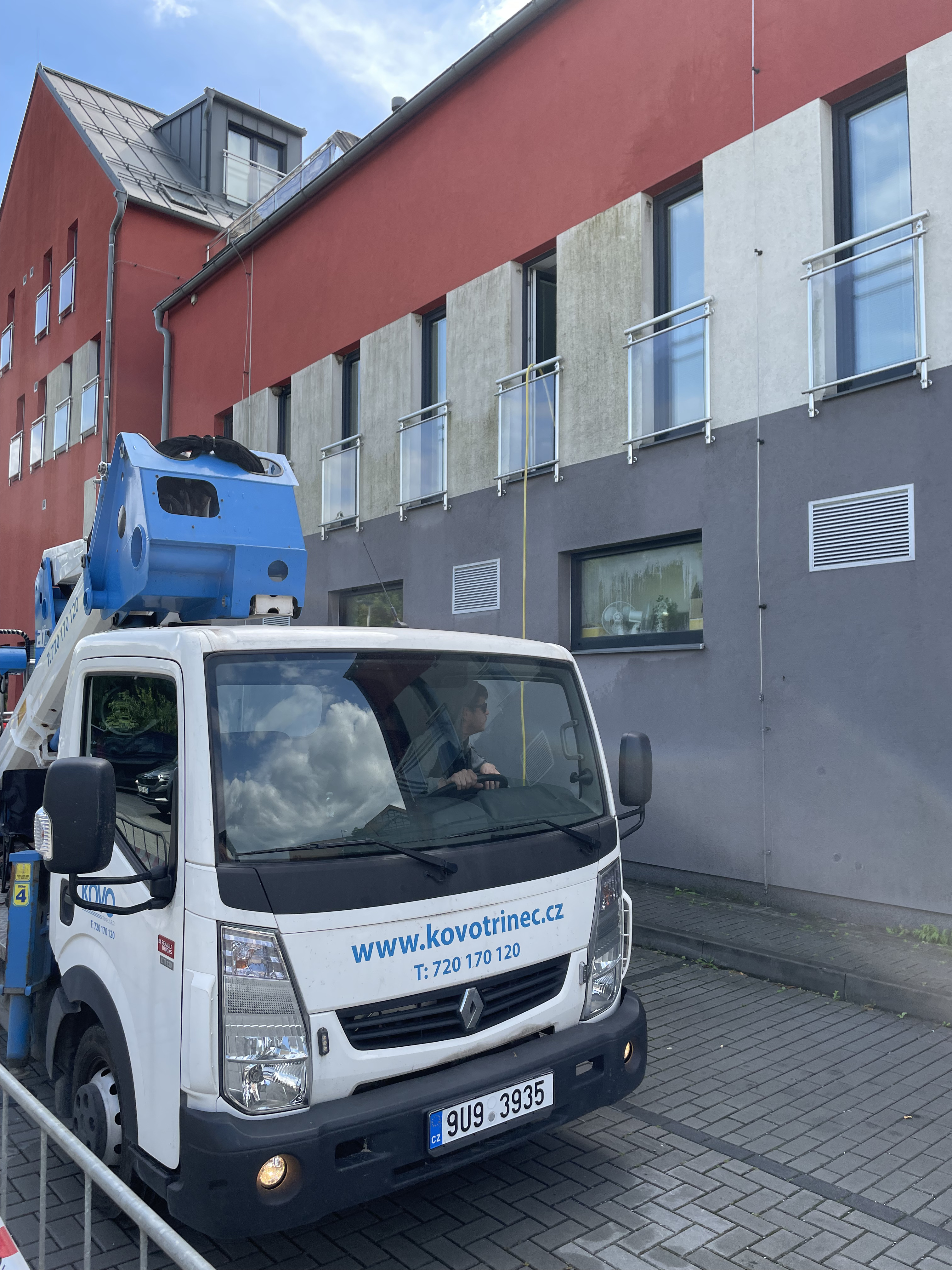 White utility truck with a blue aerial lift parked on a street beside a modern building with red, gray, and white facade.