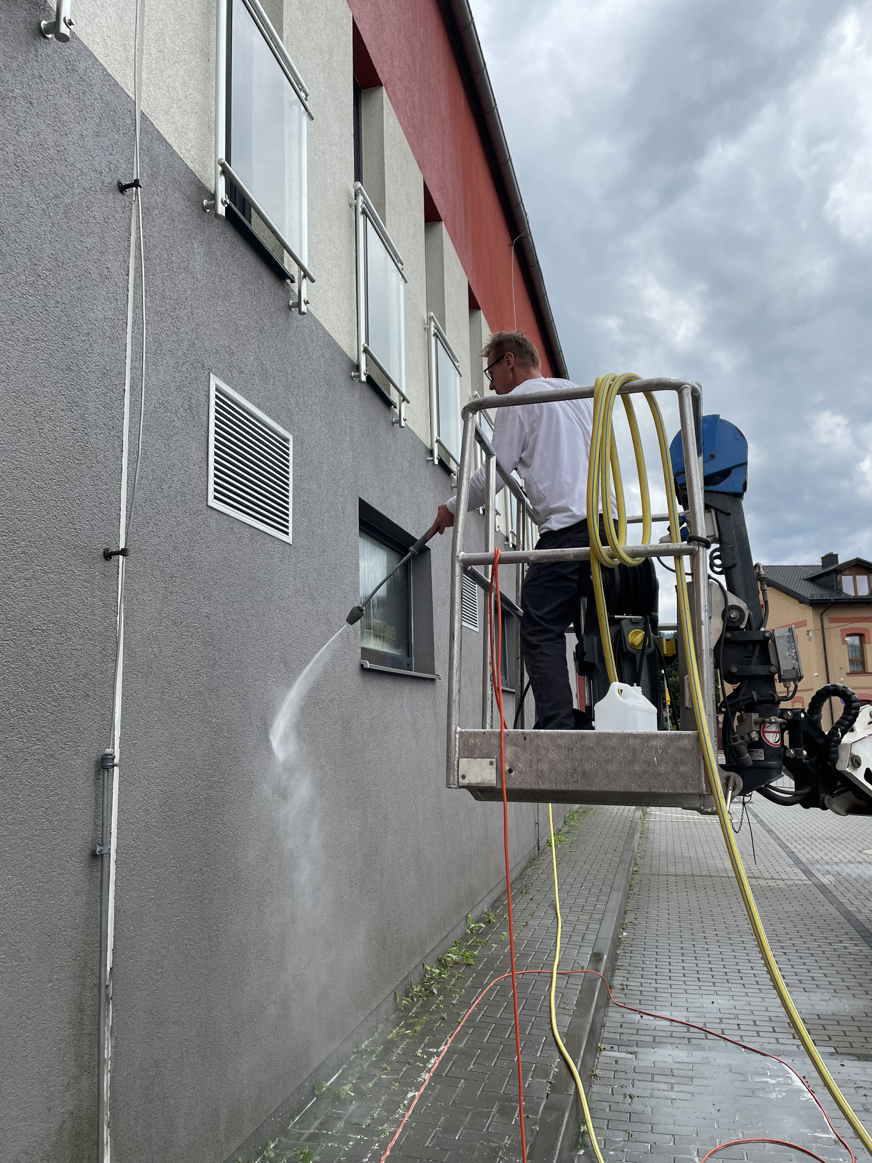Man in a white shirt pressure washing the exterior gray wall of a building while standing on a mechanical lift platform.