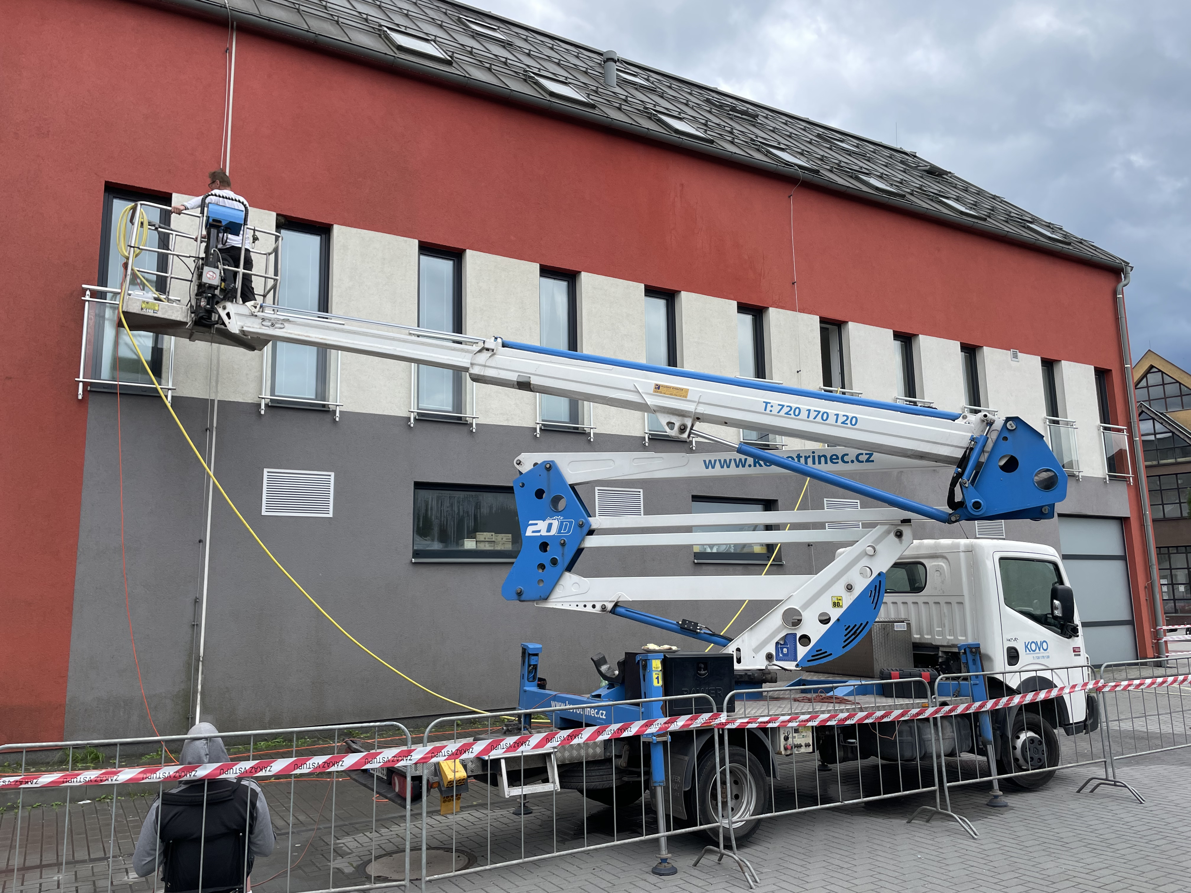 Man using a blue and white hydraulic lift truck to clean windows on a red and gray building while another person stands below behind safety barriers.