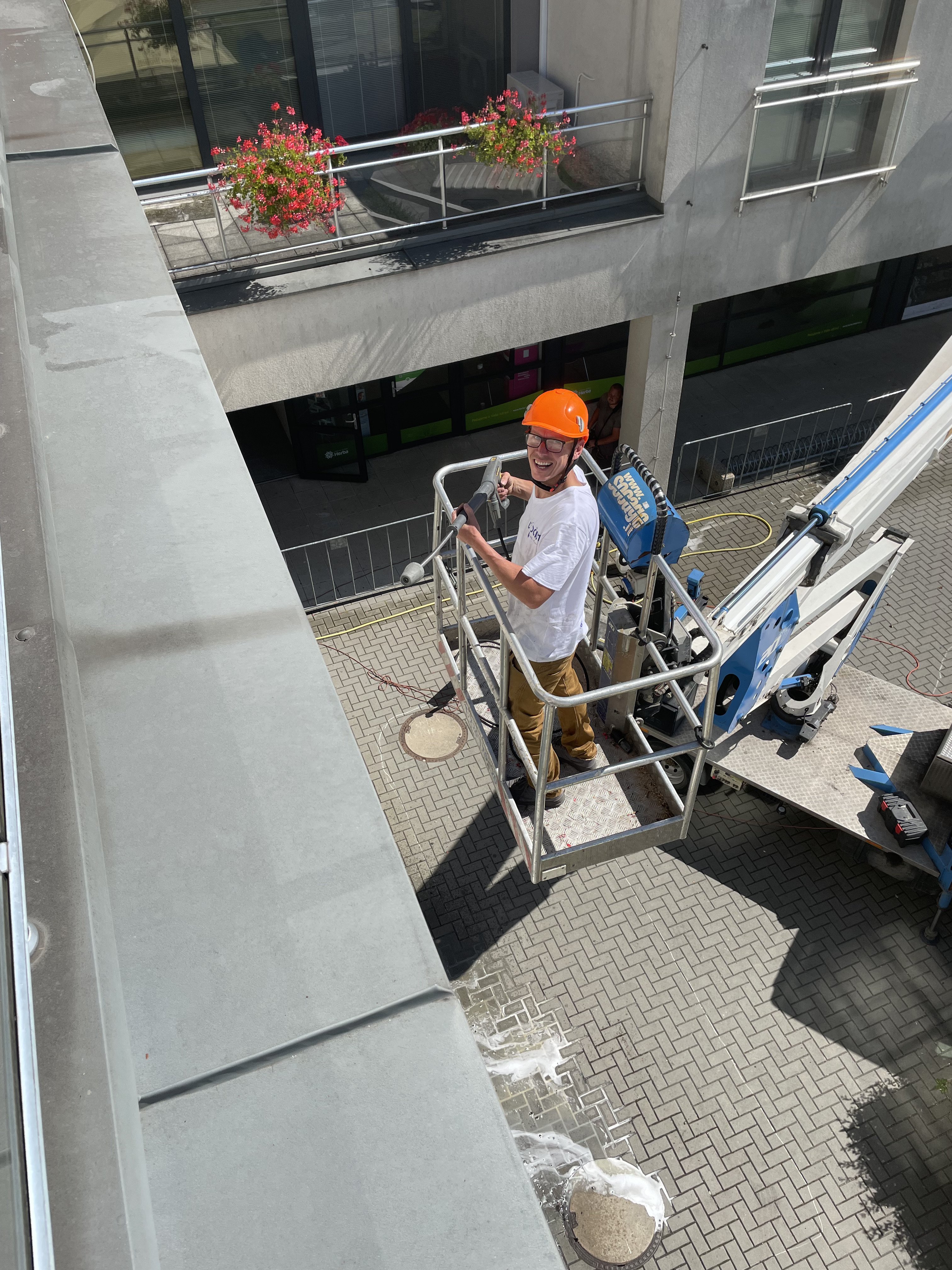 Smiling worker wearing an orange helmet operating a hydraulic lift platform near a building facade with flowers in the background.