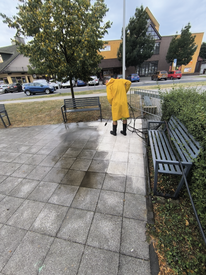 Person wearing a yellow raincoat and black boots pressure washing a tiled outdoor area, showing a clear contrast between cleaned and dirty tiles.