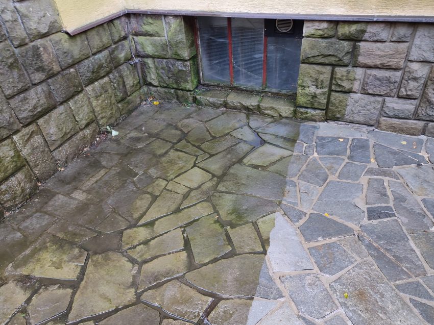 Stone-paved ground with wet and dry sections in front of a stone building wall with a small window.